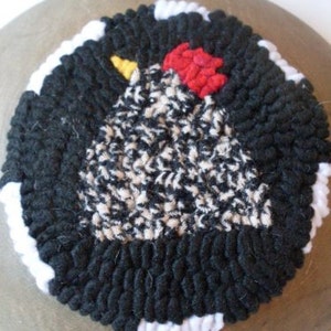 May include: A black and white rug with a rooster design. The rooster is made of white, black, and red yarn. The rug is round and has a white border.