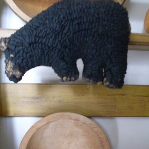 May include: A black, fuzzy, handmade animal figurine resembling a sheep or a bear. The figurine is hanging from a string and is positioned above a wooden surface.