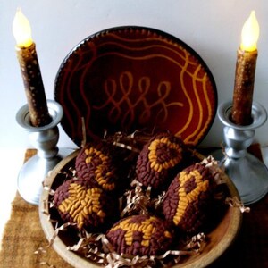 May include: Five brown and orange wool felt ornaments shaped like eggs, arranged in a wooden bowl with brown paper shreds. The bowl is on a brown surface with two lit candles in silver candle holders on either side.