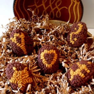 May include: Five brown and yellow felt pincushions shaped like eggs are nestled in brown shredded paper inside a brown ceramic bowl.