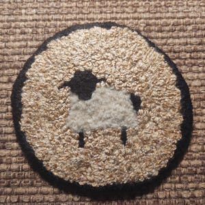 May include: A round, black-framed rug with a white sheep design in the center. The sheep is made of fluffy white yarn and has a black head and legs.