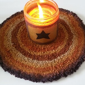 May include: A lit candle in a glass jar with a star design sits on a round, hand-hooked rug. The rug features concentric circles of brown, tan, and orange hues. The candle flame glows brightly, casting a warm light.
