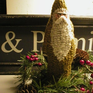 May include: A green and white felt Santa Claus figure with a white beard and a red berry wreath around the base. The wreath also includes pine cones and green pine branches.