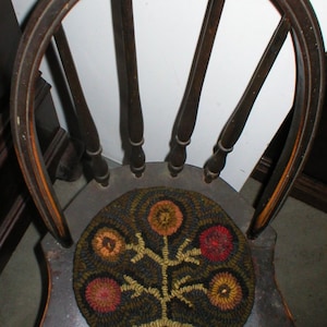 May include: A wooden chair with a dark brown finish and a round, colorful, hand-stitched rug on the seat. The rug features a tree with five flowers in shades of red, orange, and yellow.