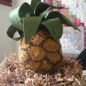 Puede incluir: Un alfiletero en forma de piña marrón y naranja con hojas de fieltro verde. El alfiletero está sentado en un cuenco marrón lleno de virutas de papel marrón.