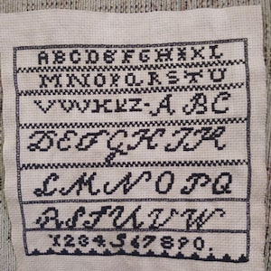May include: A white cross-stitch sampler with black embroidery of the alphabet, numbers, and punctuation marks.