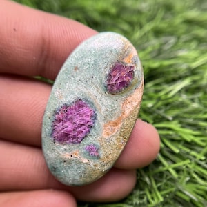 Puede incluir: Una piedra preciosa en cabujón de forma ovalada que presenta una matriz de zoisita verde pálido lisa con tres cristales de rubí magenta intenso incrustados. La superficie muestra una textura natural pulida con variaciones de color visibles.