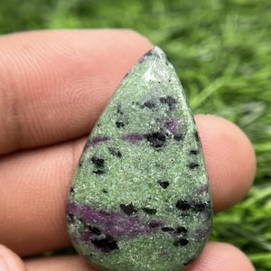 Puede incluir: Una piedra preciosa en forma de lágrima con una base de color verde claro moteado, inclusiones negras dispersas y parches de color magenta púrpura vibrante en toda la superficie pulida y texturizada.