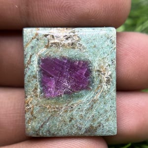 May include: A rectangular polished gemstone featuring a vibrant deep magenta center encased in a textured light green matrix. The stone displays natural mineral inclusions and a smooth, flat surface with visible crystalline patterns throughout.