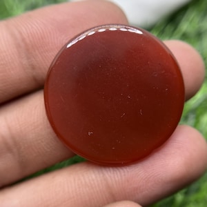 Puede incluir: Una piedra preciosa cabujón circular, lisa y pulida, de un color rojo anaranjado profundo y sólido. La piedra presenta una superficie plana y redondeada con un acabado brillante que refleja la luz, mostrando un aspecto opaco y consistente.