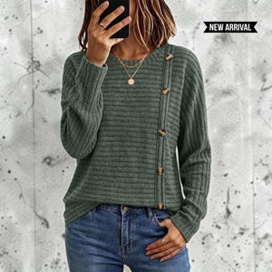 May include: A long-sleeved, ribbed knit jumper in a muted olive green colour. The jumper features a crew neckline and a vertical row of five brown buttons running down the left side of the chest. A black banner with the text NEW ARRIVAL is visible.