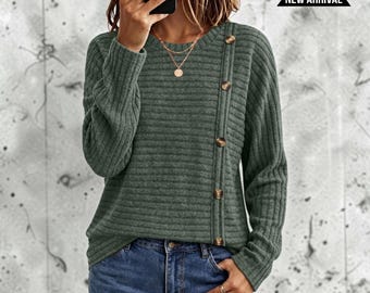 May include: A long-sleeved, ribbed knit jumper in a muted olive green colour. The jumper features a crew neckline and a vertical row of five brown buttons running down the left side of the chest. A black banner with the text NEW ARRIVAL is visible.