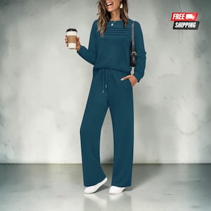 Op de afbeelding: Een blauwgroene set bestaande uit een top met lange mouwen en horizontale stiksels op de borst en een bijpassende broek met wijde pijpen en een trekkoord in de taille. De outfit wordt gecombineerd met witte sneakers en een zwarte schoudertas.