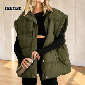 May include: An olive green sleeveless puffer vest featuring a high collar, a front zipper closure, and snap buttons. The vest has a quilted design with horizontal stitching. A black long-sleeve top is worn underneath. Text reads NEW ARRIVAL.