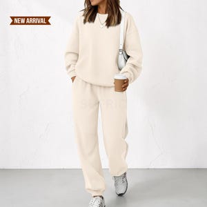 Op de afbeelding: Een crèmekleurige set bestaande uit een sweatshirt met lange mouwen en ronde hals en een bijpassende joggingbroek met elastische boorden. De outfit is effen van kleur en heeft een eenvoudig ontwerp. Een klein banner toont de tekst NEW ARRIVAL.
