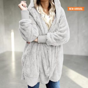 May include: A light gray hooded cardigan made of a soft, fuzzy, plush material. The garment features long sleeves with wide, ribbed knit cuffs and an open front design. An orange banner in the upper right corner displays the text NEW ARRIVAL.