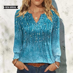 May include: A long-sleeved blouse in a vibrant teal blue with a textured, speckled finish. The garment features a V-neckline and a delicate pattern of thin, dark vertical lines with small white and yellow accents. A black tag reads NEW ARRIVAL.