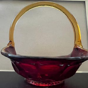 Puede incluir: Una cesta de cristal de color rojo intenso con una superficie exterior texturizada y facetada, y un asa arqueada de cristal de color ámbar. La cesta tiene una parte superior ancha y abierta con bordes ligeramente acampanados y una base circular.