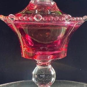 Puede incluir: Un cuenco para dulces de cristal rojo arándano con tapa a juego, que presenta un remate de bola de cristal transparente, un borde con cuentas y una base de pedestal de cristal transparente con un tallo esférico. El recipiente tiene una boca ancha.