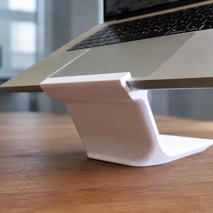 Puede incluir: Un soporte blanco e inclinado para portátil que sostiene un ordenador plateado. El soporte presenta una base lisa y curva y una superficie superior plana que sujeta el borde del equipo, elevando el teclado y la pantalla en un ángulo.