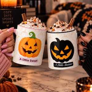May include: Two ceramic mugs filled with whipped cream and cinnamon sticks. The left mug is white with a pink handle, an orange jack-o'-lantern, and the text A little Sweet. The right mug is white with a black handle, a black jack-o'-lantern, and the text A Little Sinister.