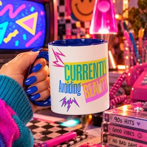 May include: A white ceramic mug with a blue interior and handle, featuring the text "CURRENTLY Avoiding REALITY" in teal, blue, and yellow, accented by pink and purple lightning bolt graphics, set against a vibrant, retro-themed background.