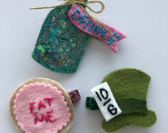 Op de afbeelding: Vilt haarclips met Alice in Wonderland-thema. Inclusief een glitterfles met "DRINK ME"-label, een roze koekje met "EAT ME"-tekst en een groene hoed met een "10/6"-label. Elke clip heeft een glitteraccent.