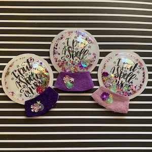 May include: Three snow globe-shaped pins with the text "I put a spell on you" in a cursive font. The globes are filled with glitter and star-shaped confetti. Each pin has a felt base in purple or lavender, with a decorative embellishment.