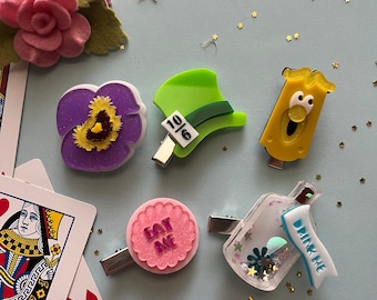 Op de afbeelding: Een verzameling haarclips met het thema Alice in Wonderland. Inclusief een roze vilten roos, een paarse viooltje, een groene Mad Hatter-hoed, een geel personage, een roze "Eat Me" clip en een "Drink Me" flesclip. Speelkaarten en gouden sterrenconfetti.