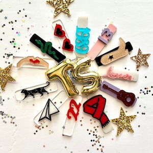 May include: A collection of colorful acrylic charms with various designs, including a snake, sunglasses, a guitar, a star, a plane, a cat, a scarf, a boot, and the letters "TS". The charms are arranged in a circle around a gold star and surrounded by gold glitter.