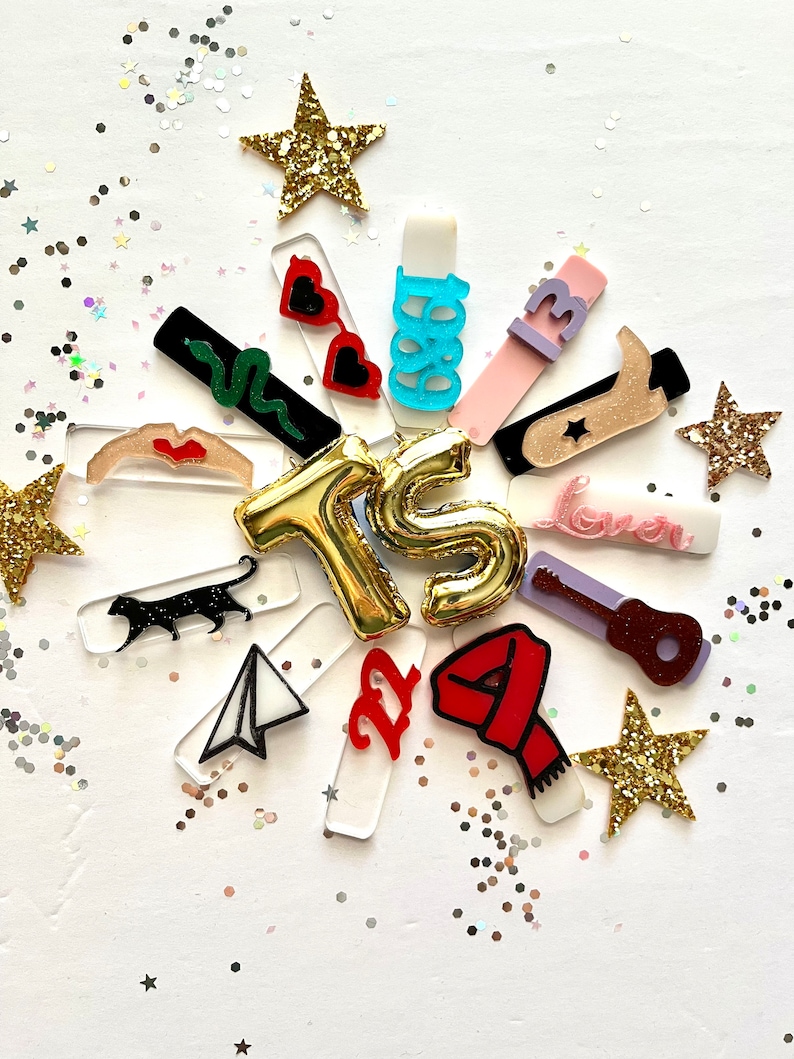 May include: A collection of colorful acrylic charms with various designs, including a snake, sunglasses, a guitar, a star, a plane, a cat, a scarf, a boot, and the letters "TS". The charms are arranged in a circle around a gold star and surrounded by gold glitter.