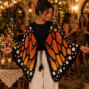 May include: A crochet butterfly wing shawl featuring a vibrant orange and yellow gradient pattern on a black background with white floral accents along the edges. The garment includes a black tie closure at the center front with two black tassels.