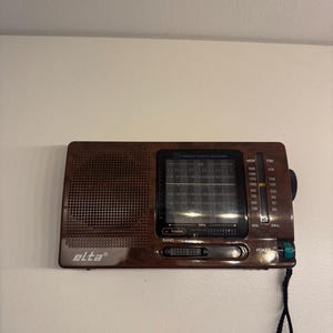 Op de afbeelding: Een bruine draagbare radio van het merk Elta met een houtnerfafwerking, voorzien van een luidsprekerrooster aan de linkerkant, een centraal frequentiedisplay voor MW, SW en FM, een afstemknop aan de rechterkant en een aan-uitknop met polsband.