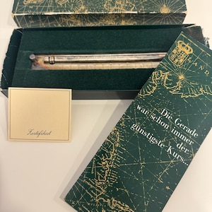 May include: A silver-toned metal ruler with engraved markings housed in a dark green rectangular box featuring a gold map design. The box lid displays the text Die Gerade war schon immer der günstigste Kurs and includes a small cream certificate card.