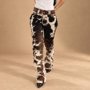 Op de afbeelding: Lange broek met een koeienprint in zwart, wit en bruin. De broek heeft een hoge taille met een knoopsluiting, riemlussen en een textuur die aanvoelt als vacht over het gehele kledingstuk, wat zorgt voor een opvallende en gedetailleerde uitstraling.