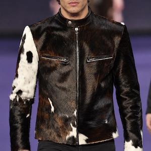 May include: A jacket featuring a cowhide pattern with dark brown, black, and white patches. The garment has a mandarin collar with a single snap button, a full-length front zipper, and two horizontal zippered chest pockets.