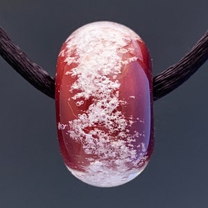 Peut inclure: Une perle de verre rouge avec des taches blanches et des bulles d'air, suspendue à un cordon noir.