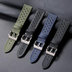 Könnte beinhalten: Vier Uhrenarmbänder in Olivgrün, Dunkelgrau, Marineblau und Schwarz. Jedes Armband hat eine strukturierte Oberfläche mit Perforationsmuster, weiße Kontrastnähte an den Rändern und eine polierte Dornschließe aus silberfarbenem Metall.