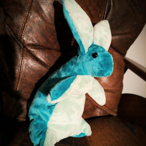 May include: A plush bunny toy in shades of teal and light blue. The stuffed animal has long ears and a soft, furry texture. The bunny is sitting on a brown leather chair.