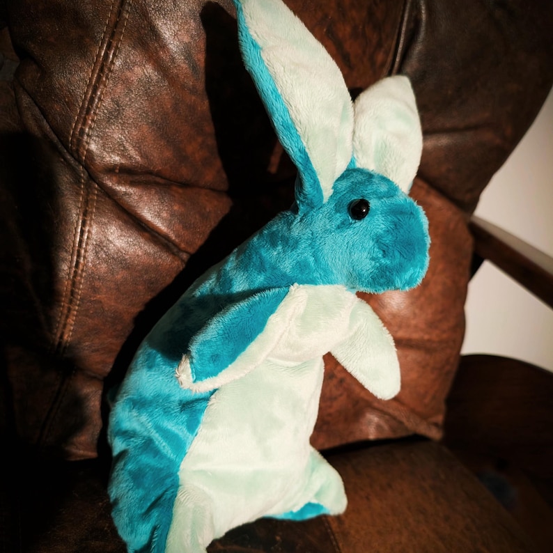 May include: A plush bunny toy in shades of teal and light blue. The stuffed animal has long ears and a soft, furry texture. The bunny is sitting on a brown leather chair.