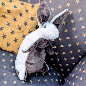 May include: A plush bunny rabbit toy with white and brown fur, long ears, and black eyes. The stuffed animal is resting on a dark gray cushion with gold star patterns, next to a yellow pillow with dark blue stars.