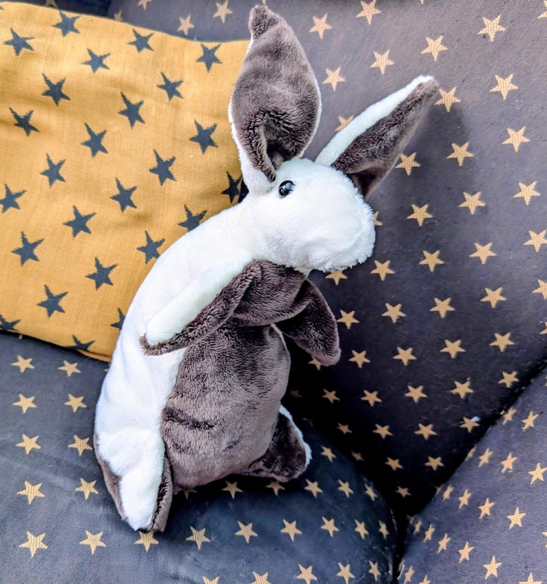 May include: A plush bunny rabbit toy with white and brown fur, long ears, and black eyes. The stuffed animal is resting on a dark gray cushion with gold star patterns, next to a yellow pillow with dark blue stars.