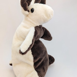 May include: A plush toy rabbit in a standing position, featuring a two-tone design. The rabbit has a cream-colored body with dark brown accents on the ears, paws, and back. It has a soft, fluffy texture and a black eye.