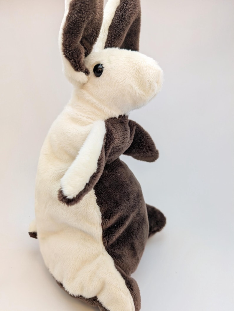 May include: A plush toy rabbit in a standing position, featuring a two-tone design. The rabbit has a cream-colored body with dark brown accents on the ears, paws, and back. It has a soft, fluffy texture and a black eye.