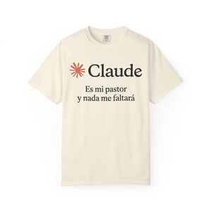 May include: A cream-colored short-sleeve t-shirt featuring a red starburst icon next to the black text 'Claude' and the Spanish phrase 'Es mi pastor y nada me faltará' printed in a serif font on the center of the chest area of the garment.