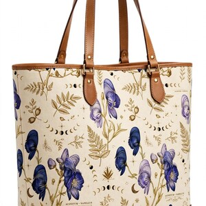 Puede incluir: Un bolso tote de color crema con un estampado botánico de flores de acónito moradas, hojas de helecho doradas y ciclos de fases lunares. El bolso incluye asas de cuero marrón, ribetes de cuero marrón y detalles de esquinas metálicas doradas.