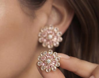 May include: A pair of floral-shaped stud earrings featuring a central clear stone surrounded by two concentric rings of marquise-cut light pink and clear stones set in a metallic frame, with one earring worn on the ear and one held in the fingers.