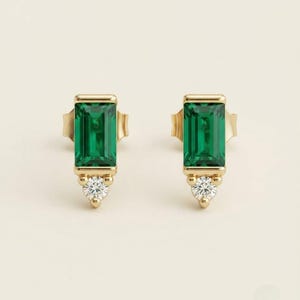 May include: A pair of gold stud earrings featuring a rectangular emerald-cut green gemstone set vertically, accented by a single small round white diamond at the base of each earring, secured with a standard post and butterfly back closure.