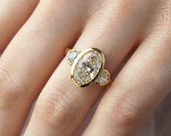 May include: A gold ring featuring a large oval-cut center stone set in a smooth bezel, flanked by two smaller round-cut stones on either side. The band is crafted from polished yellow gold, providing a warm metallic contrast to the clear, faceted stones.