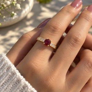 Peut inclure: Une bague en or avec une pierre précieuse rouge foncé de taille ovale au centre, entourée de trois petites pierres claires de chaque côté de l'anneau. La bande métallique présente une finition polie et les pierres sont fixées par des griffes.